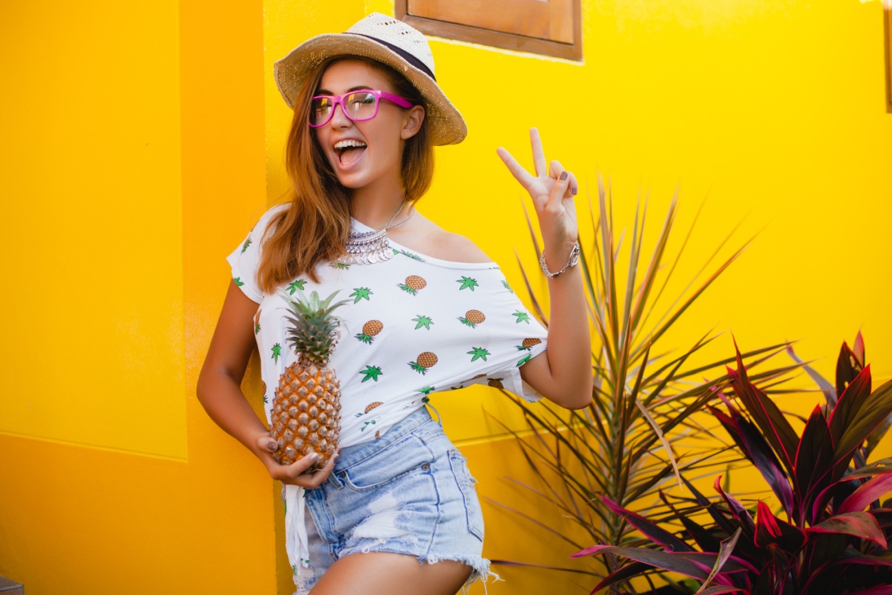 Mulher com roupas de verão sorri segurando um abacaxi. Ela usa um chapéu de palha
